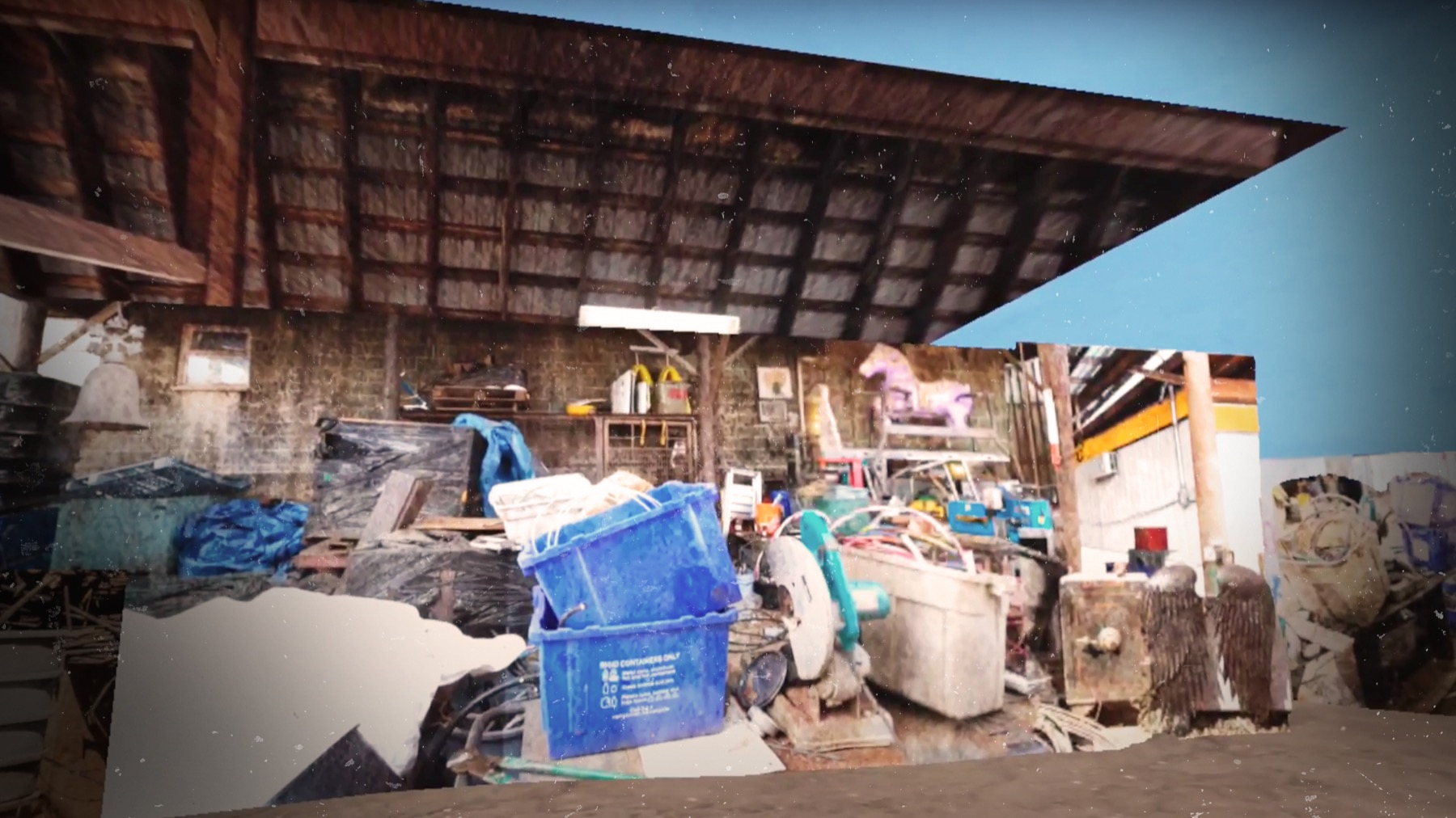 A salvage yard interior under an open roof, piled with crates, tarpaulins and discarded objects