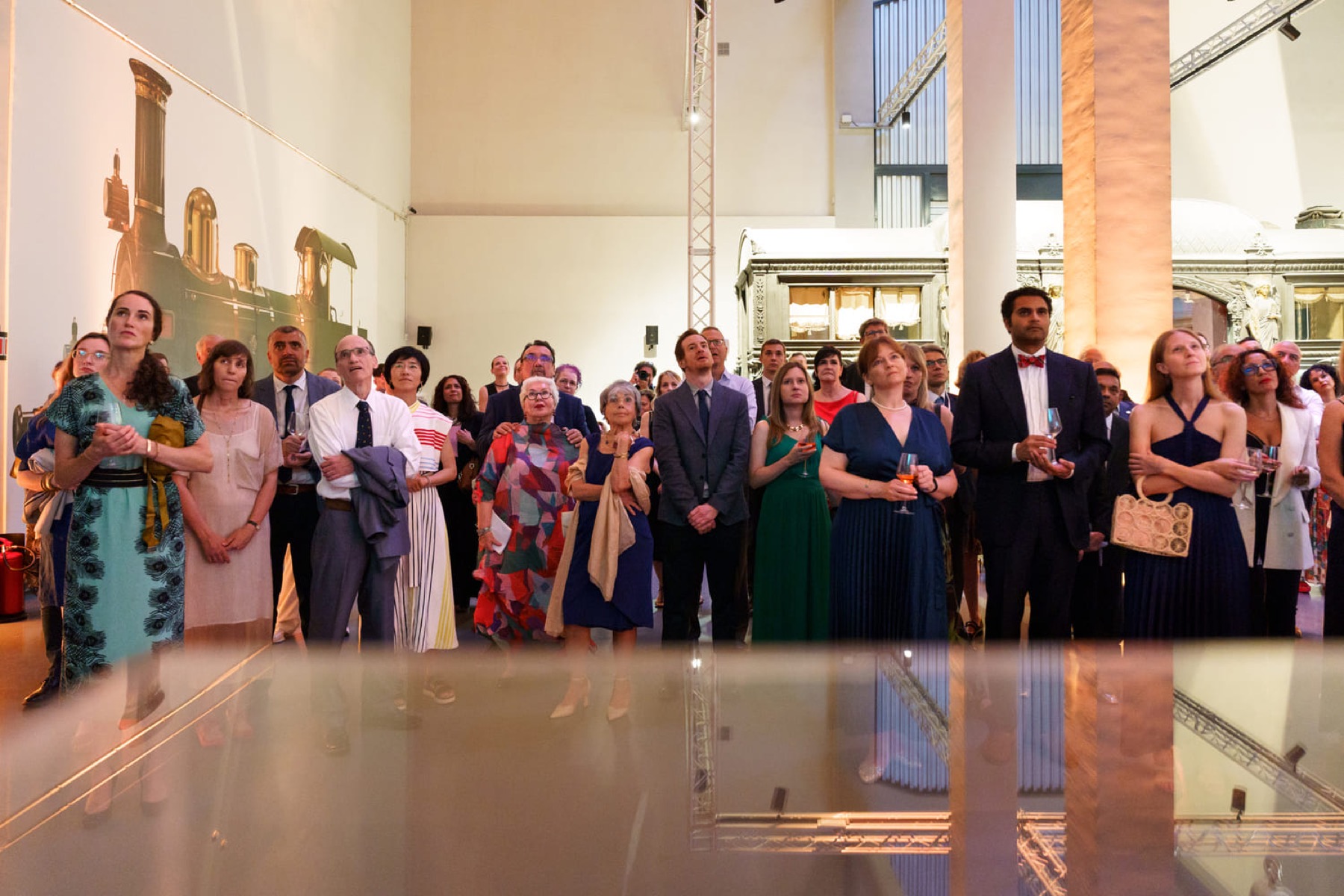 An audience standing and looking up at a screen, reflected in a polished floor