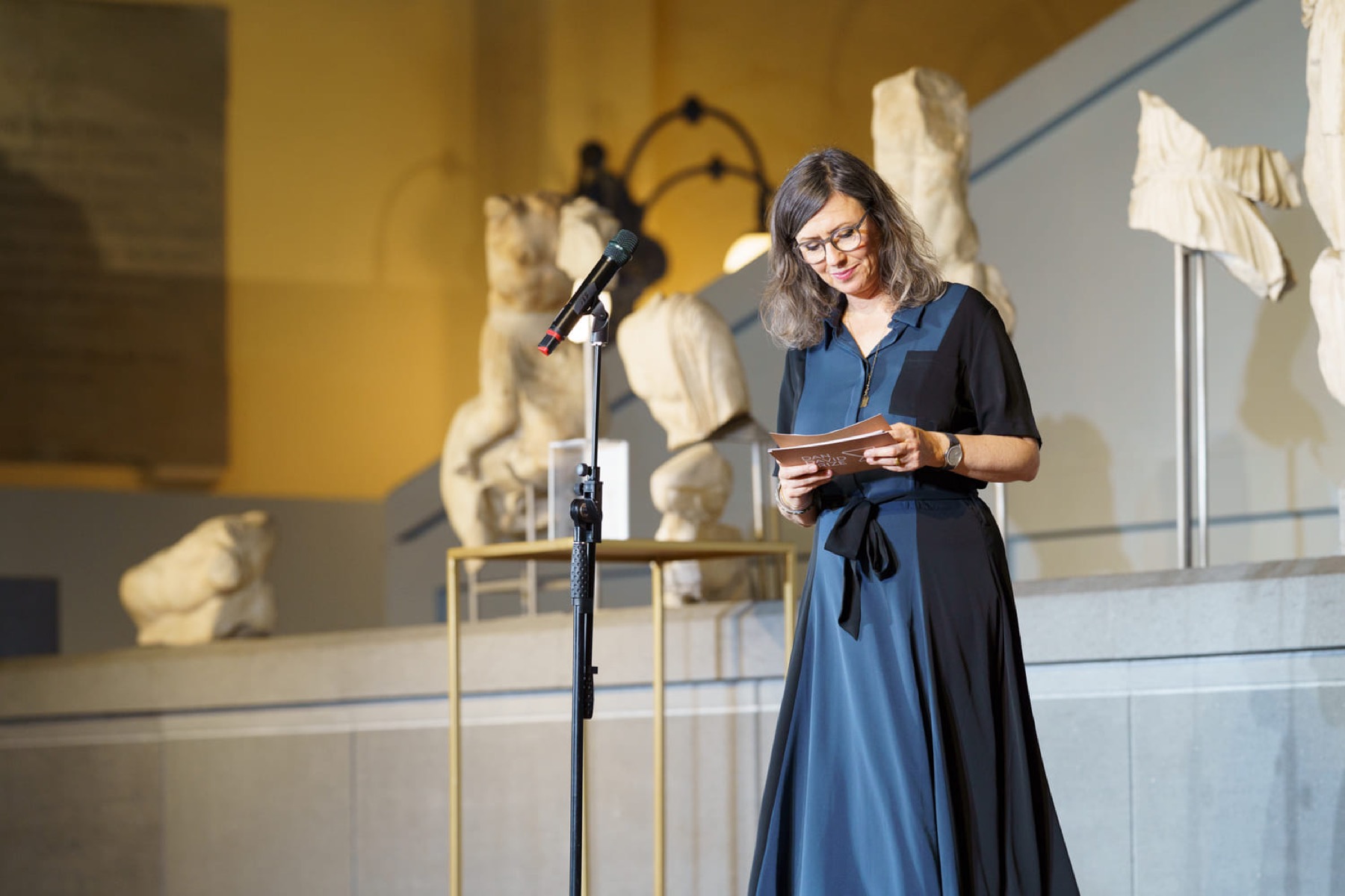 A speaker reading from a Dan David Prize programme at a microphone, classical sculpture behind her